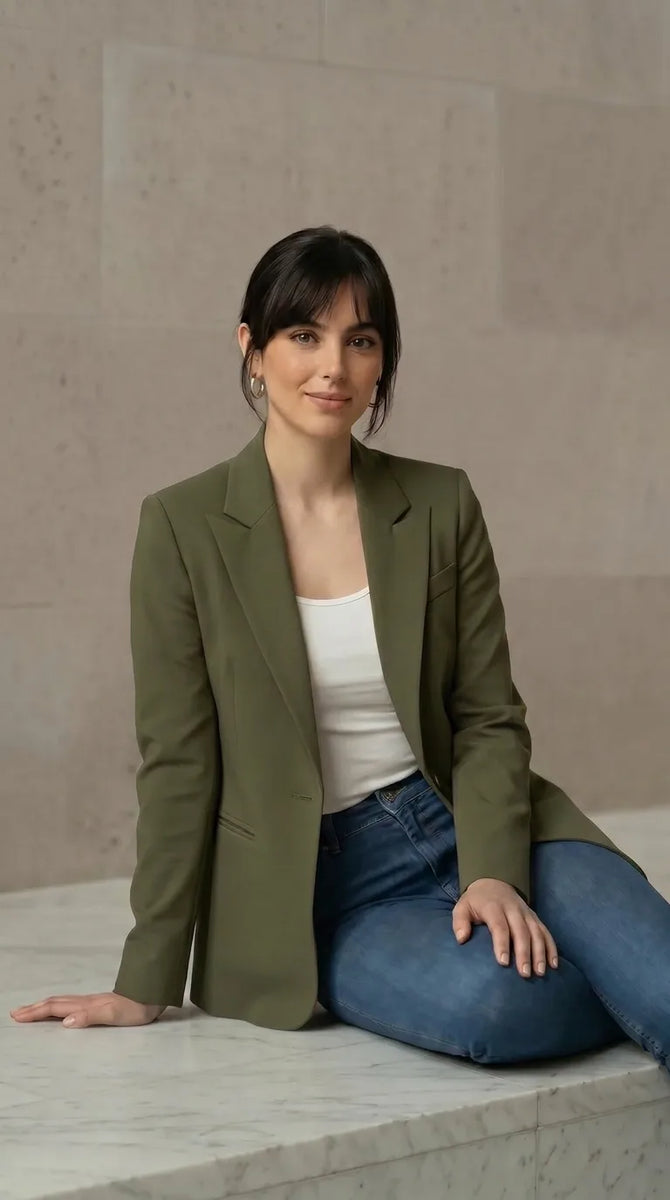 Woman wearing an olive blazer and blue jeans sitting on a marble ledge.