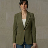 A woman wearing an olive single-breasted blazer with a gold button, styled over a white top and blue jeans, standing against a neutral stone wall.