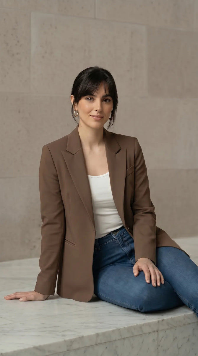 Woman wearing a mocha blazer and blue jeans sitting on a stone ledge.