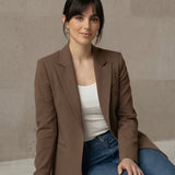 Woman wearing a mocha blazer and blue jeans sitting on a stone ledge.