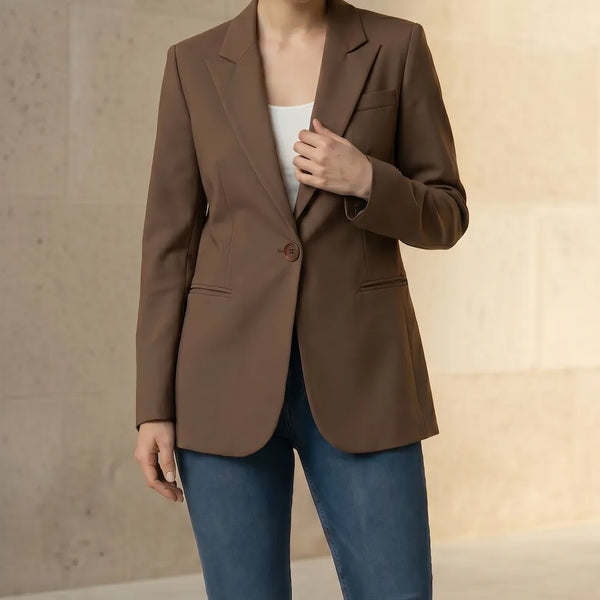 Woman wearing a mocha blazer and blue jeans with a beige handbag on a neutral background