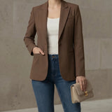 Woman wearing a mocha blazer, white top, blue jeans, and holding a beige handbag against a neutral background.
