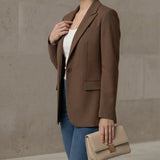 Woman wearing a mocha blazer and blue jeans, holding a beige handbag against a neutral background