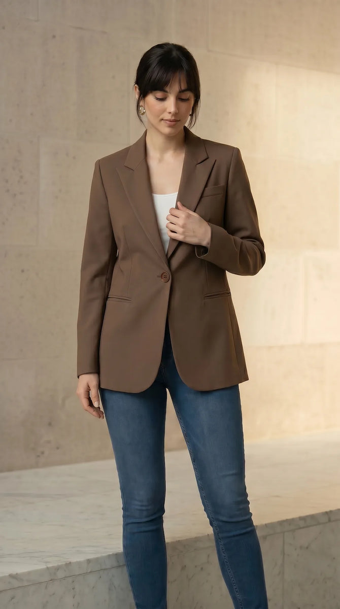 Woman wearing a mocha blazer and blue jeans against a beige wall.