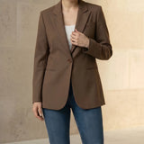 Woman wearing a mocha blazer and blue jeans against a beige wall.